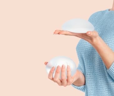 Close-up of a woman in a light blue sweater holding two breast implants, comparing different sizes and shapes against a neutral background