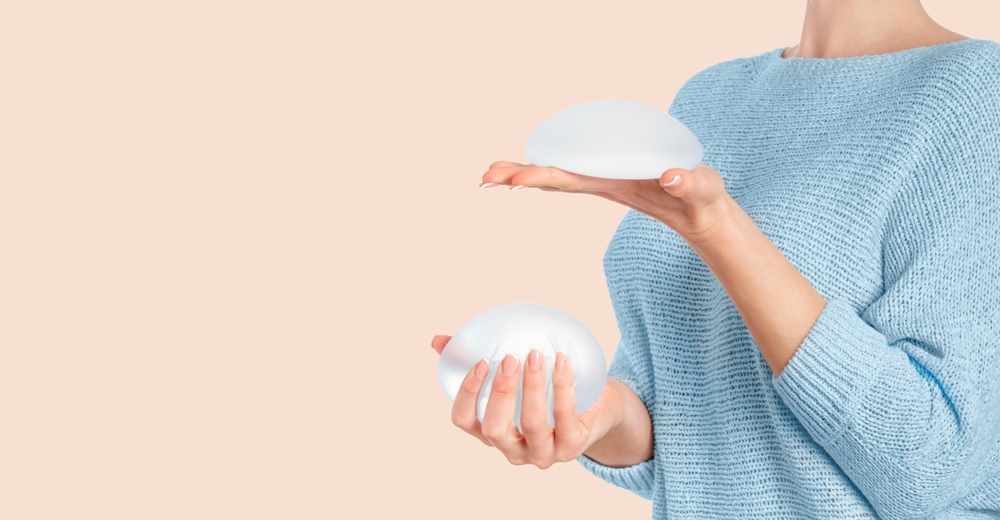 Close-up of a woman in a light blue sweater holding two breast implants, comparing different sizes and shapes against a neutral background